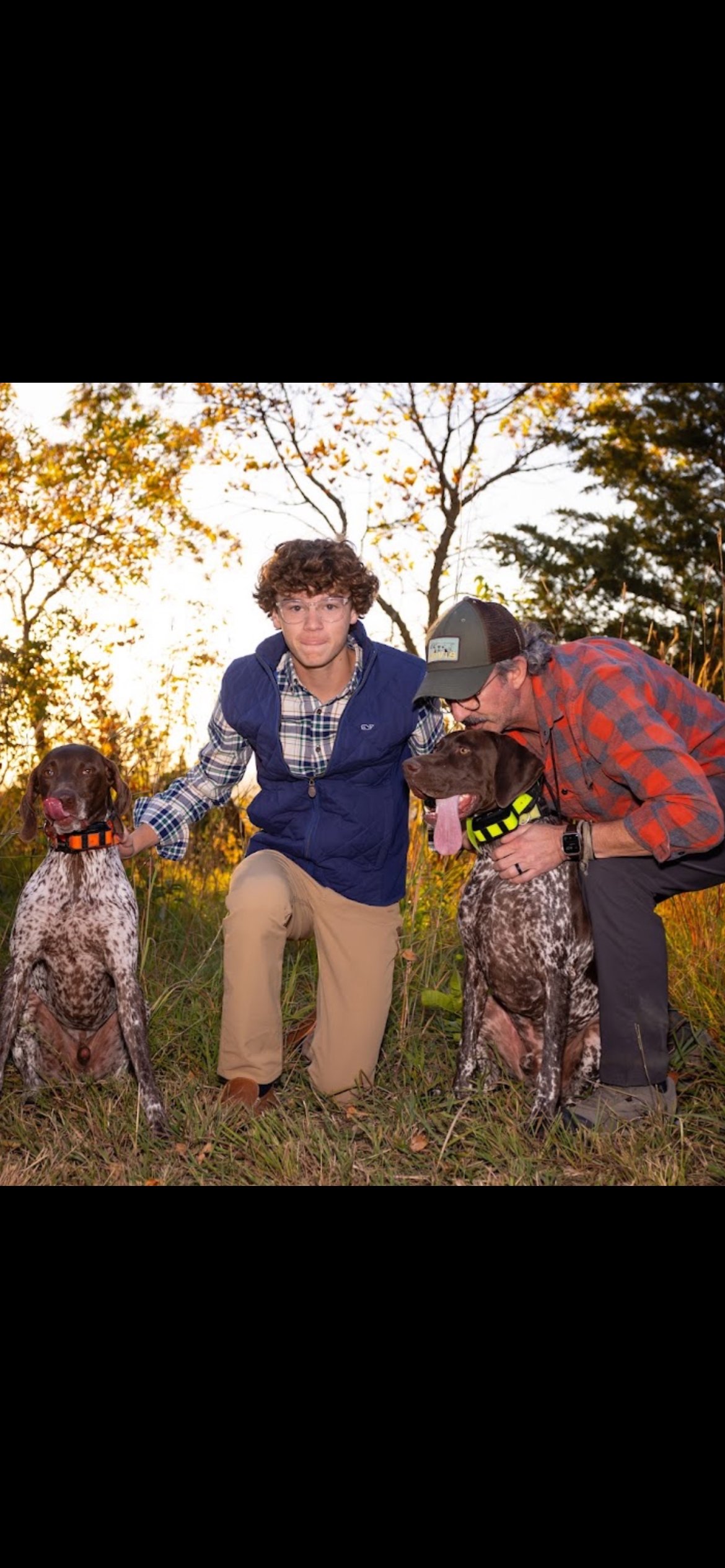 Easton and JJ Crouse kneeling in a Nebraska autumn field with Brüs in an orange collar and Karl in a green collar at golden hour.