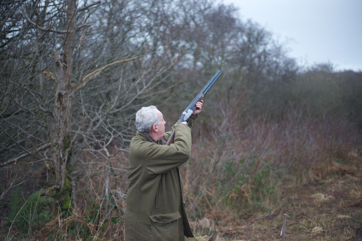 Hunter in a green shooting jacket aiming an over/under shotgun in a wooded autumn field.