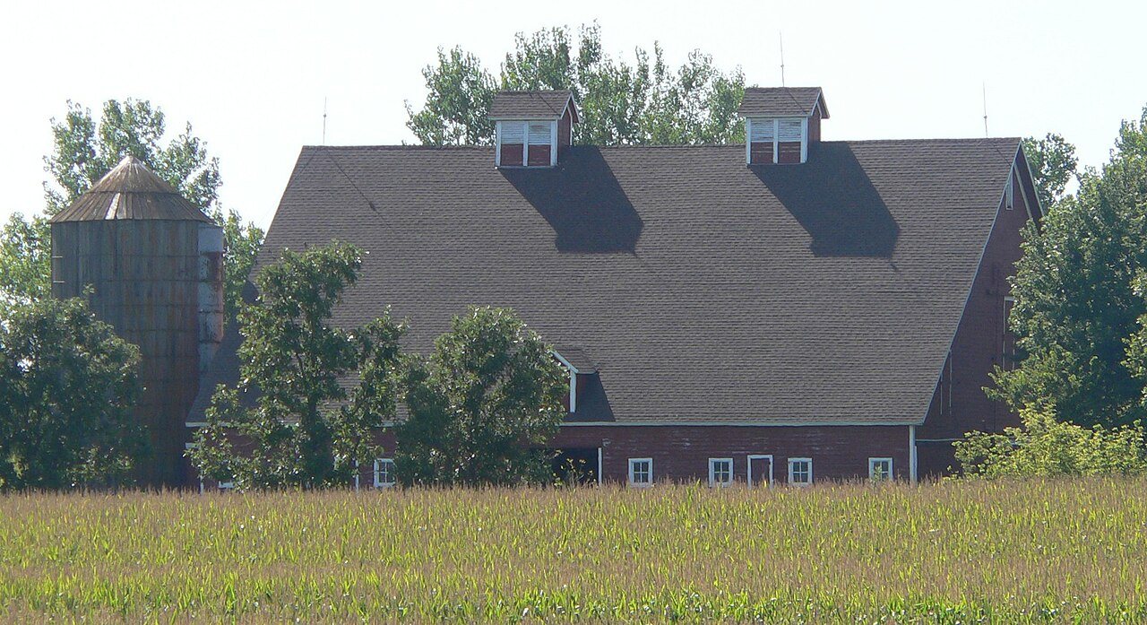 Red barn with a shingled roof and a wooden silo behind a green cornfield and shade trees on a Northern Plains farmstead.