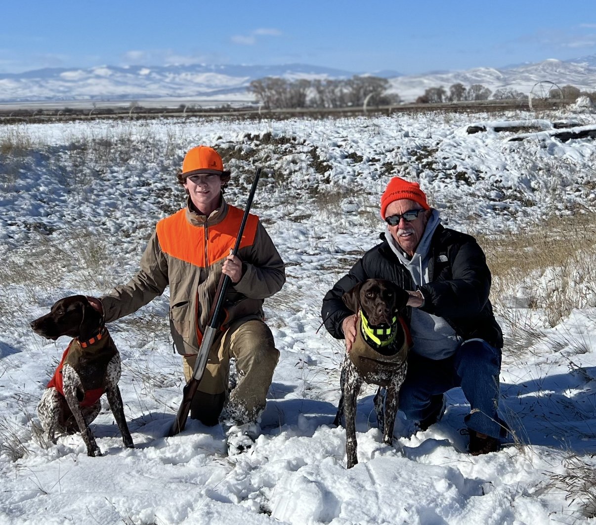 Taggart and Grandpa John kneeling in Montana snow with Brüs (orange) and Karl (green), mountains on the horizon.